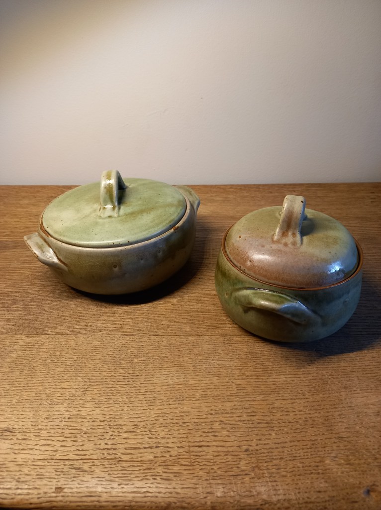 Twee keramische potten met deksels, één groen en één groen met een bruine rand, staan op een houten tafel.